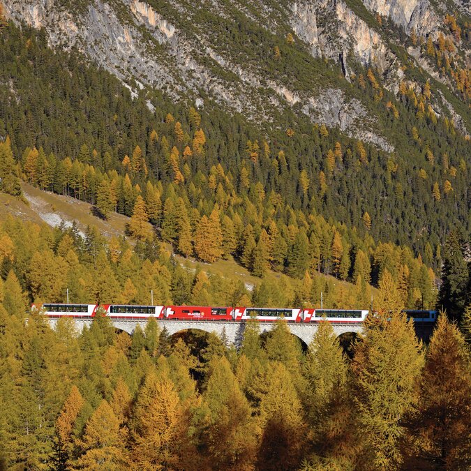 Le Glacier Express traverse un viaduc au milieu des forêts aux couleurs automnales de la vallée de l'Albula, entouré de hauts versants montagneux. | © Glacier Express